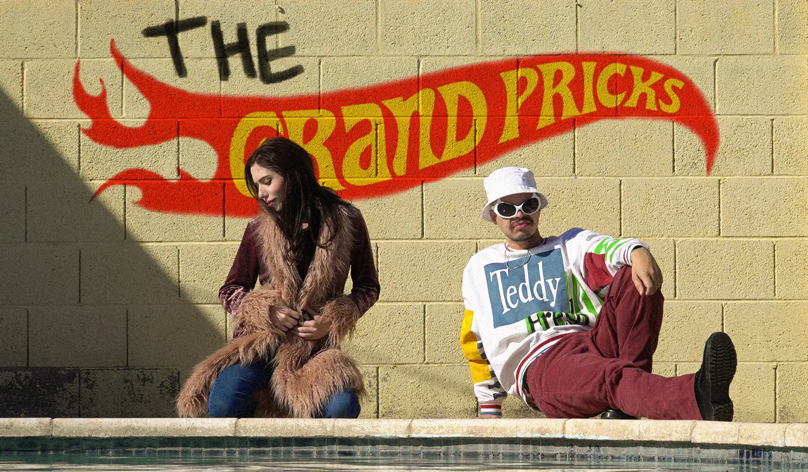 Rose Thorne and Ant Airdo of The Grand Pricks sitting by a pool under a painted Hot Wheels style Grand Pricks logo